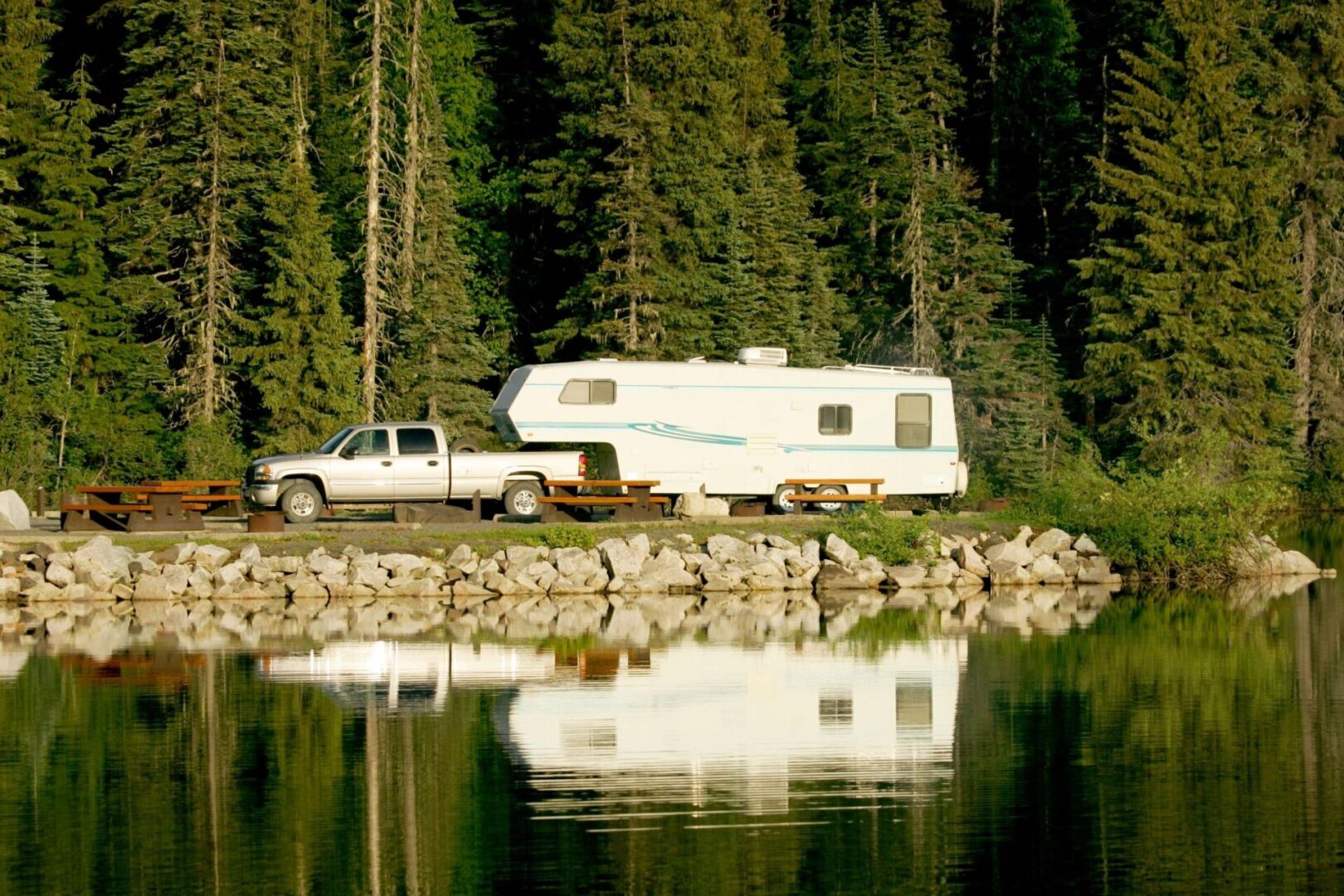 An RV parked next to a lake, surrounded by trees in the background. A truck and camper are parked on the shore of a lake.